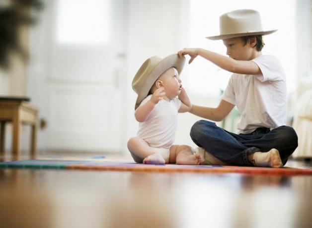 pandangan fokus selektif dari dua saudara laki-laki, satu sembilan satu delapan bulan pasangan itu duduk bersila di lantai anak laki-laki yang lebih tua meletakkan topi yang serasi di kepala bayi