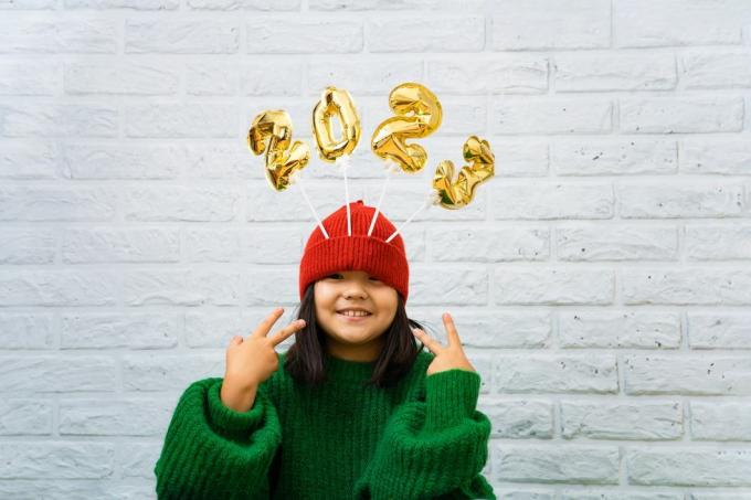 bayi perempuan asia yang bahagia dengan sweter hijau menutup dengan balon emas dengan nomor 2023 di ruang salinan topi, latar belakang dinding bata putih, gadis melihat ke kamera, tersenyum