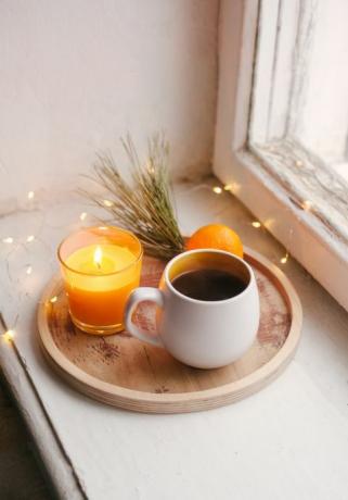 detail interior rumah musim dingin yang nyaman, secangkir kopi, nampan kayu, jeruk keprok dan ambang jendela lilin masih hidup untuk Natal