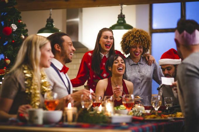 sekelompok teman tertawa ketika mereka duduk mengelilingi meja makan dan minum pada hari natal seorang pria sedang membaca lelucon dari kue natal topi pesta dan jumper natal dikenakan