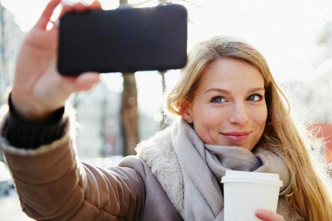 wanita tampan berjalan di jalanan kota di musim dingin mengambil selfie dan memegang kopi untuk pergi, dengan cahaya latar