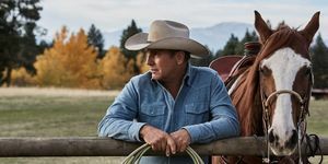 kevin costner di yellowstone di sebelah kuda bersandar di pagar dengan tali di tangan mengenakan kemeja denim biru pudar dan topi koboi krem