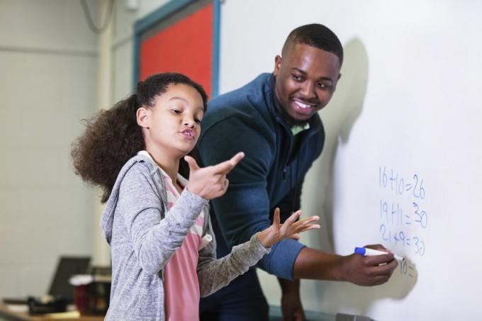 seorang gadis multiras berusia 9 tahun di kelas matematika, berdiri di papan tulis bersama gurunya, seorang pemuda Afrika-Amerika di kelasnya Di usia 20-an, guru sedang memandangi siswa SDnya, menunggu sementara dia mencoba mencari tahu jawaban yang menjadi fokus perhatiannya gadis