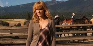 kelly reilly sebagai beth dutton dalam gaun kotak-kotak coklat putih dan merah dengan kardigan coklat berdiri di depan kandang kuda